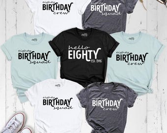 May include: A collection of six short-sleeve t-shirts in white, black, dark gray, and light blue. The shirts feature printed text including 'eighty BIRTHDAY crew', 'eighty BIRTHDAY squad', and 'hello EIGHTY est. 1946' in various black and white fonts.