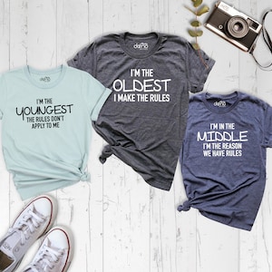 May include: Three t-shirts with different slogans about sibling order. The light blue shirt says "I'm the youngest, the rules don't apply to me". The gray shirt says "I'm the oldest, I make the rules". The dark blue shirt says "I'm in the middle, I'm the reason we have rules".