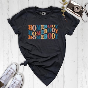 Może przedstawiać: Czarny T-shirt z napisem "HOMEBODY" powt&oacute;rzonym trzy razy w retro czcionce. Tekst jest w odcieniach pomarańczowego, czerwonego, niebieskiego i ż&oacute;łtego.