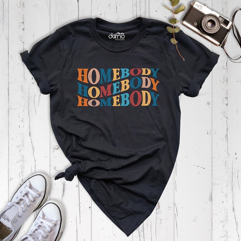Może przedstawiać: Czarny T-shirt z napisem "HOMEBODY" powt&oacute;rzonym trzy razy w retro czcionce. Tekst jest w odcieniach pomarańczowego, czerwonego, niebieskiego i ż&oacute;łtego.