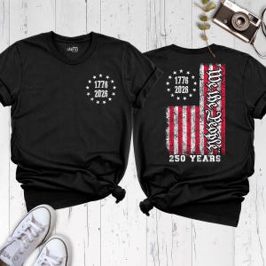 May include: A black t-shirt featuring a small white circular emblem on the chest with the text 1776 2026 surrounded by stars. The back displays a distressed American flag graphic with the text We the People and 250 YEARS in white.