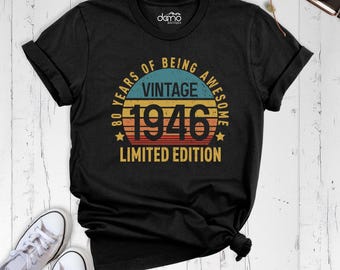 May include: A black short-sleeve t-shirt featuring a circular graphic with horizontal stripes in yellow, orange, and blue. The text reads VINTAGE 1946 80 YEARS OF BEING AWESOME LIMITED EDITION. The shirt has a crew neckline and short sleeves.