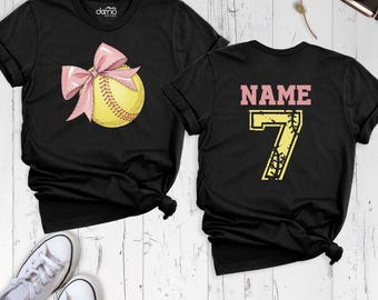 May include: A black short-sleeve t-shirt featuring a yellow softball with a pink bow on the front. The back of the shirt displays the text NAME in pink above the large yellow number 7, which is styled with a softball texture and stitching.