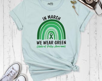 May include: A light blue t-shirt featuring a green rainbow graphic with a central awareness ribbon. The text reads IN MARCH WE WEAR GREEN Cerebral Palsy Awareness. The shirt has short sleeves and a crew neckline, with a small logo at the inner neck.
