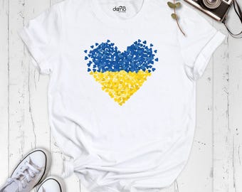 Pode incluir: Uma t-shirt branca de manga curta com um gráfico grande em forma de coração composto por muitos corações pequenos azuis e amarelos. A peça tem gola redonda e o texto "damo boutique" impresso a preto no interior da gola.