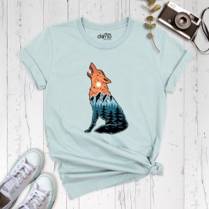 Puede incluir: Una camiseta azul claro con un gr&aacute;fico de un lobo aullando con una cordillera y una puesta de sol dentro de su silueta. El texto "domo" est&aacute; impreso en la camiseta.