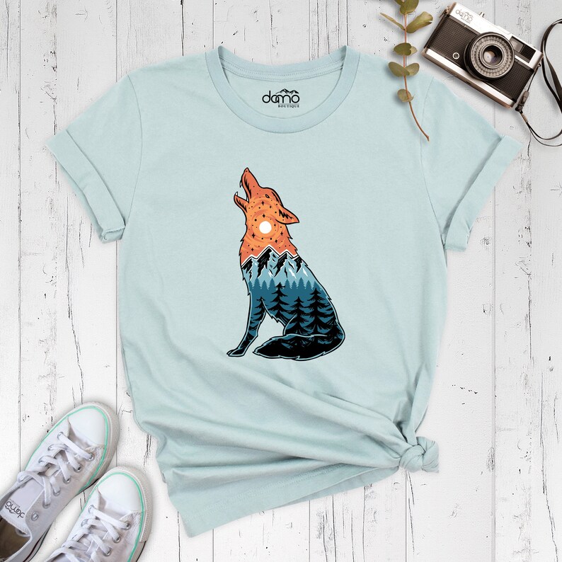 Puede incluir: Una camiseta azul claro con un gr&aacute;fico de un lobo aullando con una cordillera y una puesta de sol dentro de su silueta. El texto "domo" est&aacute; impreso en la camiseta.