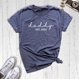 May include: A dark blue t-shirt with the text "daddy est. 2025" in white.