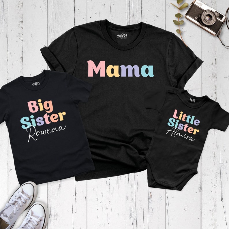 以下が含まれることがあります： 虹色の文字が書かれた黒いTシャツが3枚。一番大きいTシャツには「ママ」と書いてある。中くらいのTシャツには「ビッグシスターロウェナ」と書いてある。一番小さいTシャツには「リトルシスターアミラ」と書いてある。