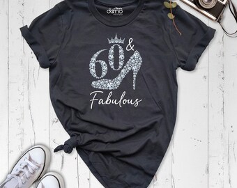 May include: A dark gray crew neck t-shirt featuring a white graphic of the number 60 and a high-heeled shoe, both filled with a diamond pattern, topped with a crown. The text "Fabulous" is printed below in white script, with "damo boutique" above.