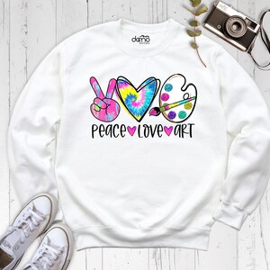May include: A white long-sleeve sweatshirt featuring a graphic print with a pink peace sign, a tie-dye heart, and an artist palette with a paintbrush. The text below reads PEACE LOVE ART with pink hearts separating the words.