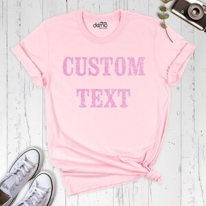 以下が含まれることがあります： 胸元にグリッターピンクのフォントで「CUSTOM TEXT」という文字がプリントされた、ライトピンクの半袖Tシャツ。丸首で、襟の内側に小さなロゴがプリントされています。生地は無地のライトピンクです。