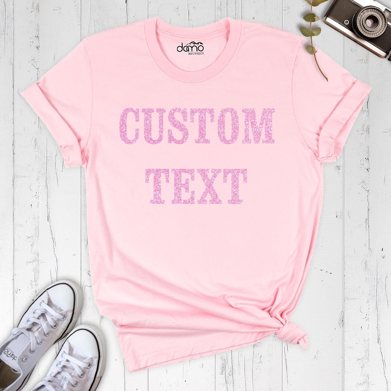 以下が含まれることがあります： 胸元にグリッターピンクのフォントで「CUSTOM TEXT」という文字がプリントされた、ライトピンクの半袖Tシャツ。丸首で、襟の内側に小さなロゴがプリントされています。生地は無地のライトピンクです。