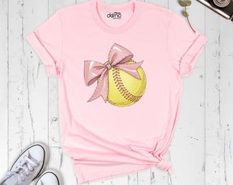 May include: A light pink short-sleeve t-shirt featuring a graphic of a yellow softball with red stitching and a large pink bow tied around it. The shirt has a crew neckline and the text "damo boutique" printed in black on the inner neck label area.