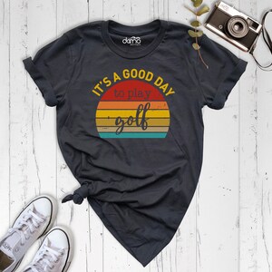 May include: A dark gray t-shirt with a vintage-style sunset graphic and the text "It's a good day to play golf".
