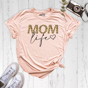 Może przedstawiać: R&oacute;żowy t-shirt z napisem "MOM life" w czarnych literach. "MOM" jest w nadruku w panterkę.