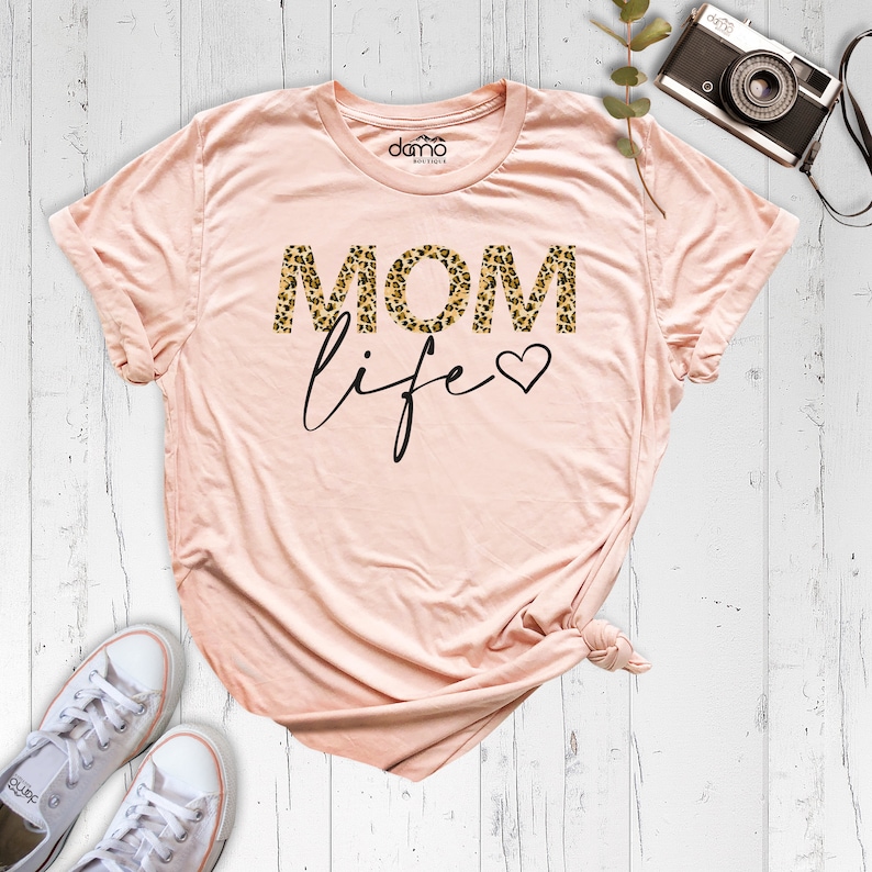 Może przedstawiać: R&oacute;żowy t-shirt z napisem "MOM life" w czarnych literach. "MOM" jest w nadruku w panterkę.