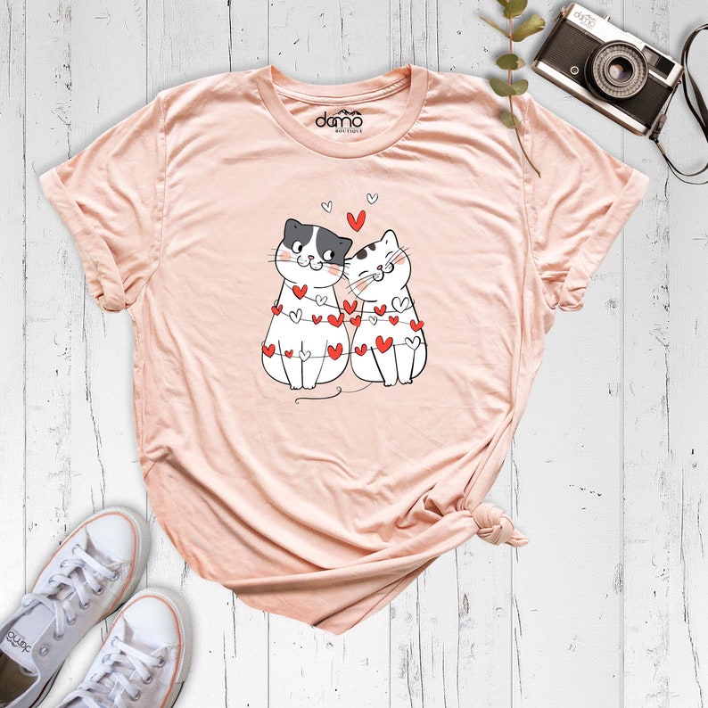 May include: A pink t-shirt with a graphic of two cats wearing a string of hearts. The text "damo boutique" is printed on the shirt.