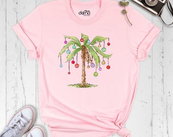 May include: A light pink short-sleeve t-shirt featuring a graphic of a palm tree decorated with colorful hanging ornaments. The shirt has a round neckline and the text "damo boutique" printed in black on the inner collar area.