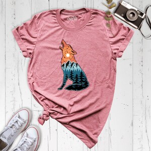 Puede incluir: Una camiseta rosa con un gr&aacute;fico de un lobo aullando a la luna. El lobo est&aacute; formado por un paisaje de monta&ntilde;a con &aacute;rboles y una puesta de sol.