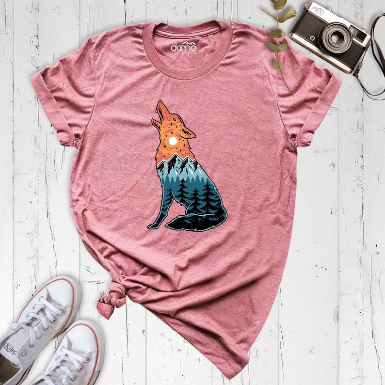 Puede incluir: Una camiseta rosa con un gr&aacute;fico de un lobo aullando a la luna. El lobo est&aacute; formado por un paisaje de monta&ntilde;a con &aacute;rboles y una puesta de sol.