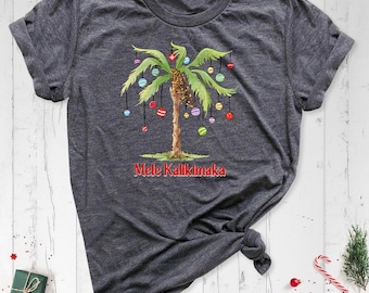 May include: A dark heather gray short-sleeve t-shirt featuring a graphic of a palm tree decorated with colorful hanging ornaments. The text Mele Kalikimaka is printed in red below the tree. The shirt has a crew neckline and a brand label.