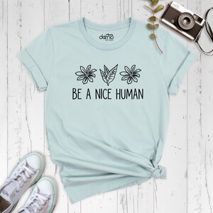 Op de afbeelding: Een lichtblauw t-shirt met een zwarte afbeelding van bloemen en bladeren en de tekst "BE A NICE HUMAN".