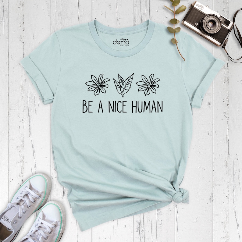 Op de afbeelding: Een lichtblauw t-shirt met een zwarte afbeelding van bloemen en bladeren en de tekst "BE A NICE HUMAN".