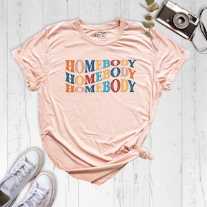Może przedstawiać: R&oacute;żowy t-shirt z napisem "HOMEBODY" powt&oacute;rzonym cztery razy w retro czcionce. Tekst jest w odcieniach niebieskiego, pomarańczowego, ż&oacute;łtego i czerwonego.
