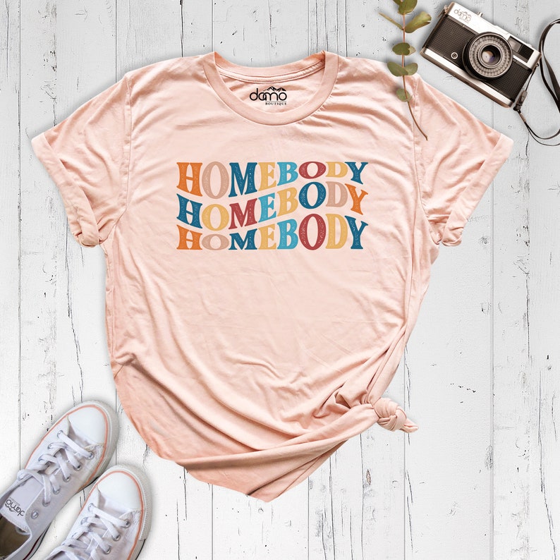 Może przedstawiać: R&oacute;żowy t-shirt z napisem "HOMEBODY" powt&oacute;rzonym cztery razy w retro czcionce. Tekst jest w odcieniach niebieskiego, pomarańczowego, ż&oacute;łtego i czerwonego.