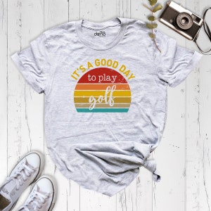 May include: A gray t-shirt with a vintage-style graphic that says "It's a good day to play golf". The graphic features a sunset with a yellow, orange, and red color scheme.
