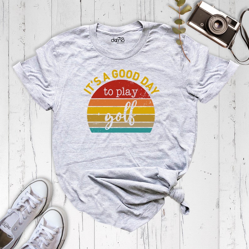 May include: A gray t-shirt with a vintage-style graphic that says "It's a good day to play golf". The graphic features a sunset with a yellow, orange, and red color scheme.
