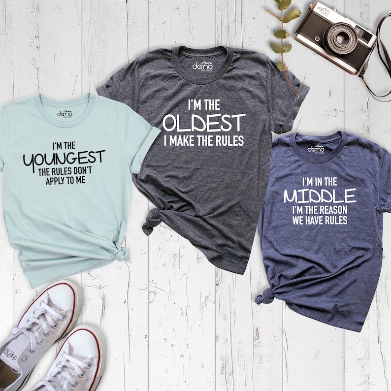 May include: Three t-shirts with different slogans about being the oldest, middle, and youngest child. The oldest shirt is gray, the middle shirt is blue, and the youngest shirt is light blue.