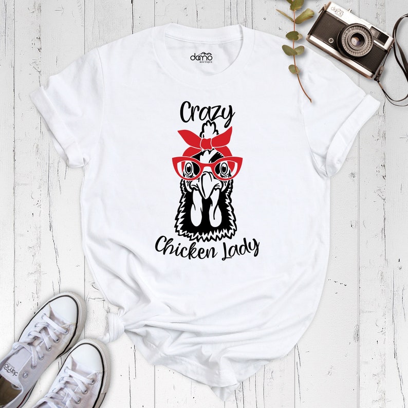 K&ouml;nnte beinhalten: Wei&szlig;es T-Shirt mit einer Schwarz-Wei&szlig;-Illustration eines Huhns mit roter Brille und rotem Bandana. Der Text "Crazy Chicken Lady" ist unter der Illustration aufgedruckt.