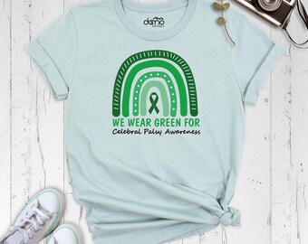 May include: A light blue t-shirt featuring a green rainbow graphic with a central awareness ribbon. Below the rainbow, the text reads WE WEAR GREEN FOR Cerebral Palsy Awareness. The shirt has short sleeves and a crew neckline.