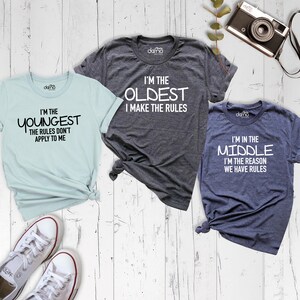 May include: Three t-shirts with different slogans about sibling order. The light blue shirt says "I'm the youngest, the rules don't apply to me". The gray shirt says "I'm the oldest, I make the rules". The dark blue shirt says "I'm in the middle, I'm the reason we have rules".