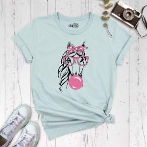 Puede incluir: Una camiseta azul claro con un gr&aacute;fico de un caballo que lleva gafas rosas y un pa&ntilde;uelo rosa con lunares blancos. El caballo est&aacute; soplando una burbuja rosa.