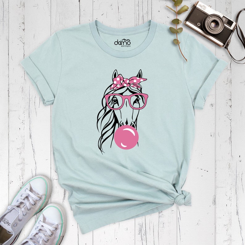 Puede incluir: Una camiseta azul claro con un gr&aacute;fico de un caballo que lleva gafas rosas y un pa&ntilde;uelo rosa con lunares blancos. El caballo est&aacute; soplando una burbuja rosa.