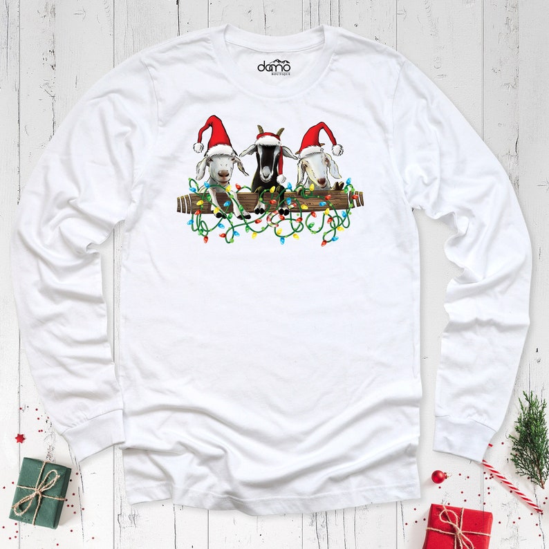 K&ouml;nnte beinhalten: Wei&szlig;es Langarmshirt mit einem Aufdruck von drei Ziegen mit Weihnachtsm&uuml;tzen, die auf einem Holzbalken sitzen, der mit bunten Weihnachtslichtern geschm&uuml;ckt ist. Das Shirt hat einen Rundhalsausschnitt und ein Markenlogo.