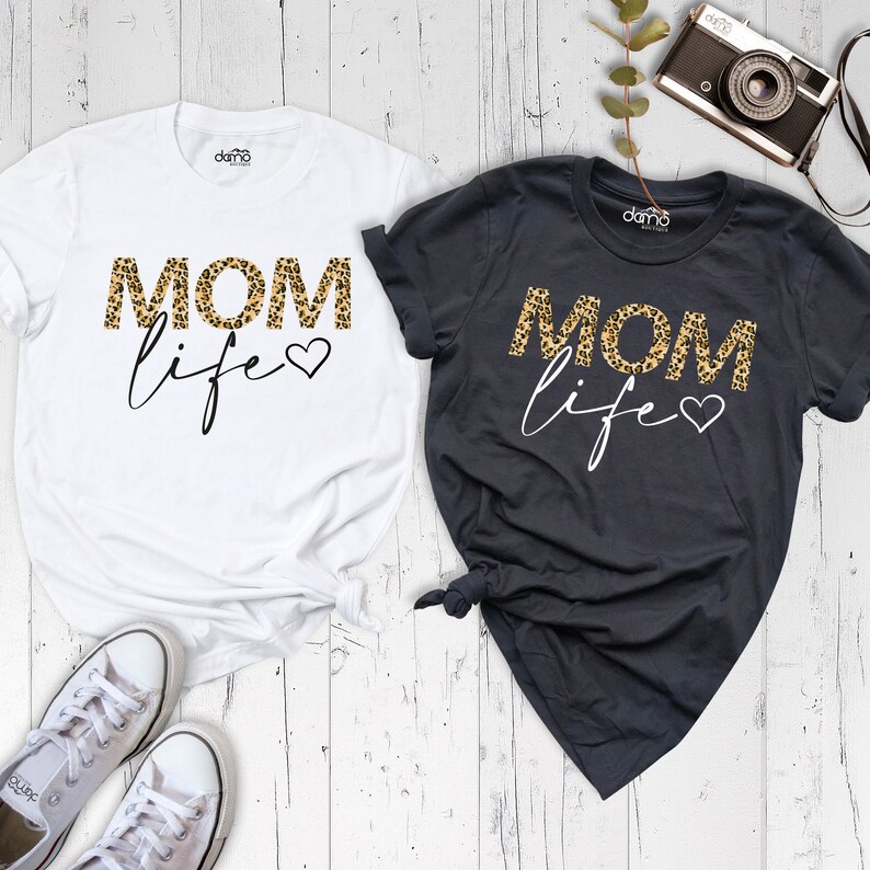 Może przedstawiać: Dwie koszulki z napisem "MOM life" w nadruku w panterkę. Jedna koszulka jest biała, a druga ciemnoszara.