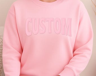 May include: A light pink crewneck sweatshirt featuring the word CUSTOM embossed in a matching pink tone across the chest. The garment has long sleeves with ribbed cuffs and a ribbed neckline. The logo Damo Boutique is visible in the top right corner.