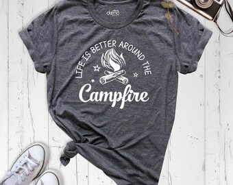 May include: A dark heather gray short-sleeve t-shirt featuring a white graphic of a campfire with logs and flames. The text reads "LIFE IS BETTER AROUND THE Campfire" in a mix of block and script fonts, accented by small stars.