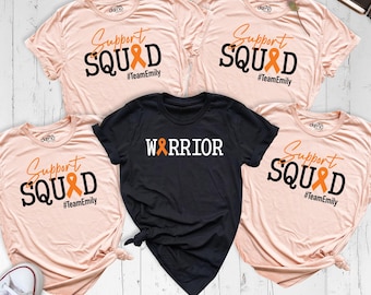 May include: Four light peach t-shirts and one black t-shirt. The peach shirts feature the text Support SQUAD with an orange ribbon replacing the letter A and the hashtag #TeamEmily. The black shirt features the text WARRIOR with an orange ribbon.
