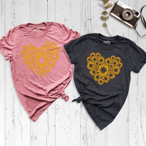 May include: Two t-shirts with a sunflower heart design. The shirt on the left is pink and the shirt on the right is dark gray.