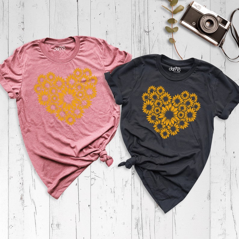 May include: Two t-shirts with a sunflower heart design. The shirt on the left is pink and the shirt on the right is dark gray.