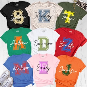 May include: A collection of short-sleeved t-shirts in various colors, each featuring a different name and initial in a collegiate style. The shirts are black, beige, green, white, red, blue, pink, and orange. The names include Santoria, Kennedy, Tanya, Andrea, Davison, Zanilo, Medison, Emily, and Jennifer.