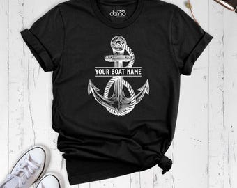 May include: A black short-sleeve t-shirt featuring a white graphic of a nautical anchor wrapped in rope. The text "YOUR BOAT NAME" is printed in white across the center of the anchor. The inner neck label displays the text "damo boutique".