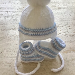 May include: White knitted baby hat with a blue and grey striped band and matching booties. The hat has a white pom pom on top.