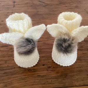 May include: A pair of white knitted baby booties with bunny ears and a gray fur pom-pom on each bootie.