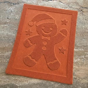 May include: A rectangular orange knitted blanket featuring a raised texture design of a gingerbread person wearing a Santa hat and a bow tie, surrounded by three small stars. The knit pattern creates a distinct embossed effect.